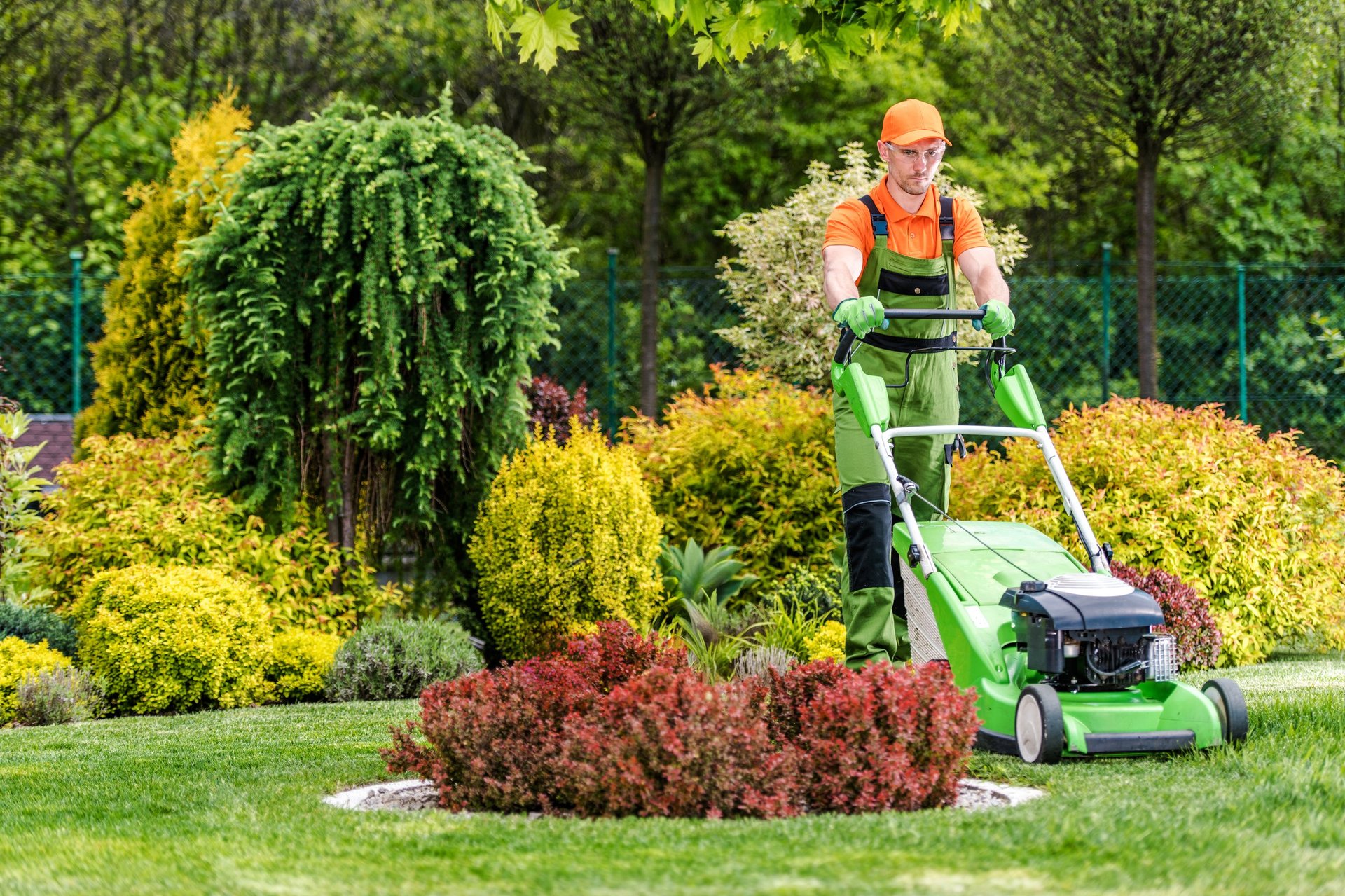 A lawn care worker is mowing grass in a garden with various plants. He wears gloves and a cap.