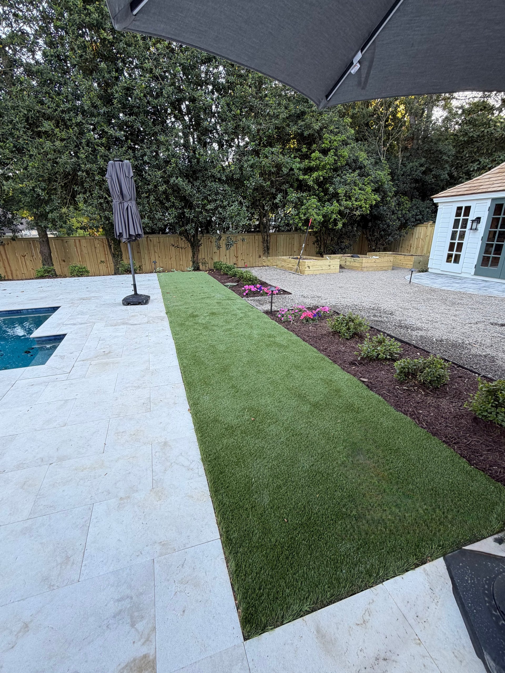 Modern backyard with artificial turf, paved patio, pool, umbrella, and landscaping beds along wooden fence