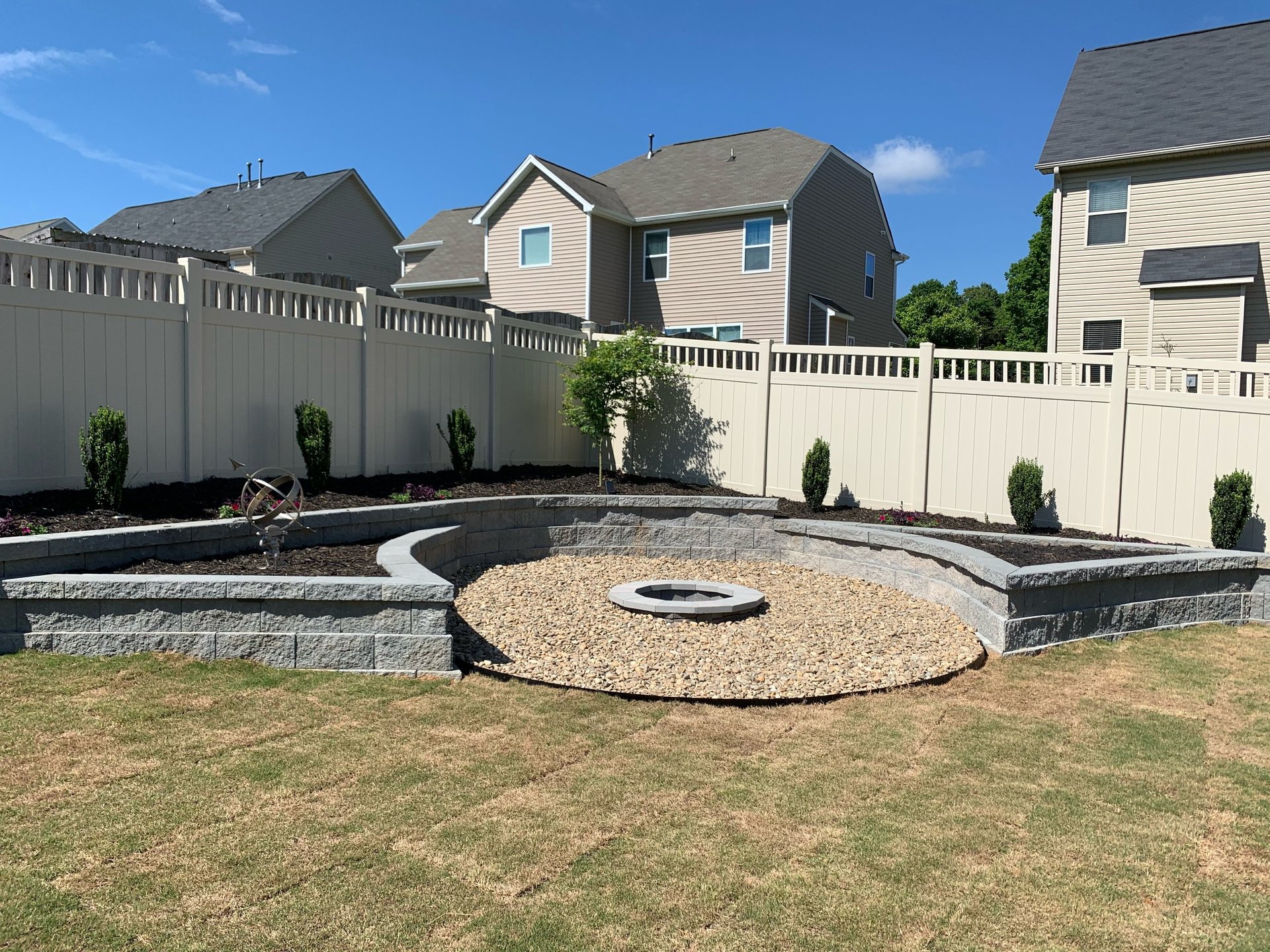 Backyard fire pit area with gravel circular design, stone retaining walls, white vinyl fencing, and landscaping with residential houses in background
