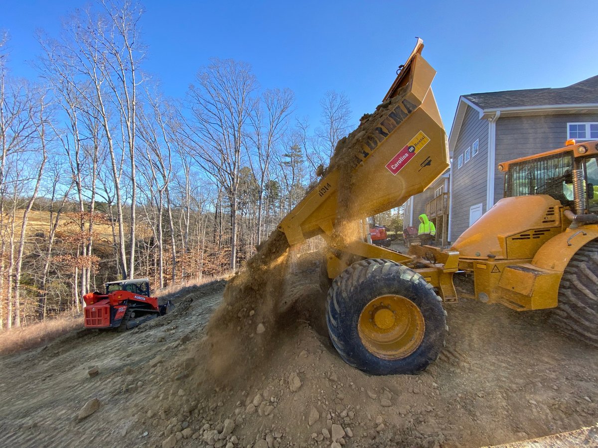 Yellow wheel loader dumping dirt and gravel at a construction site with bare trees and a house in background