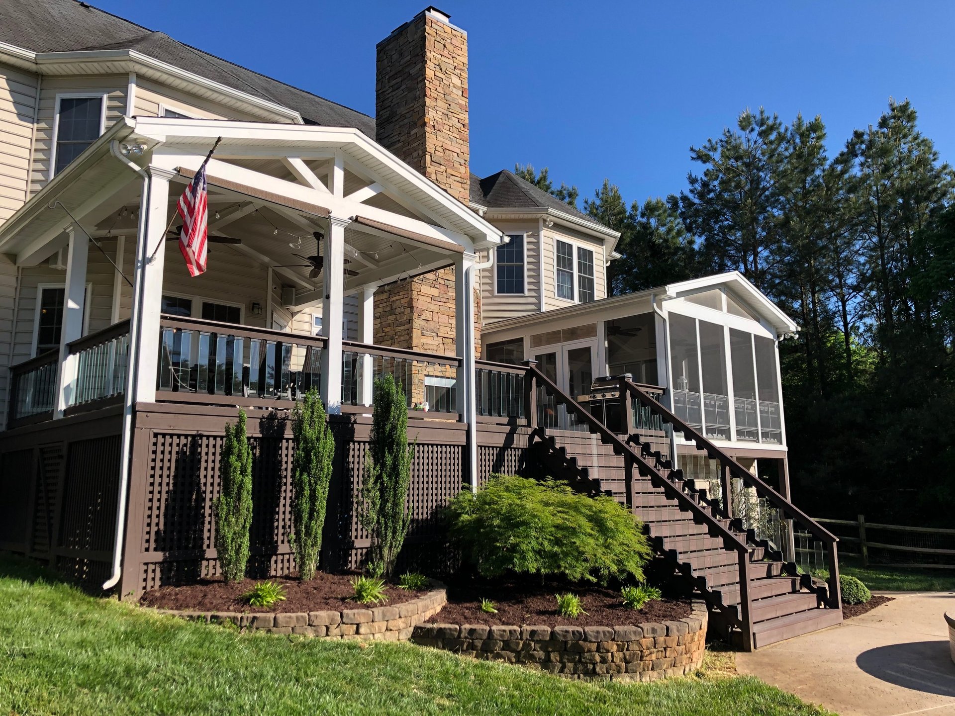 Two-story residential home with brick chimney, white pergola, metal deck railings, landscaping with shrubs, and American flag on a sunny day.
