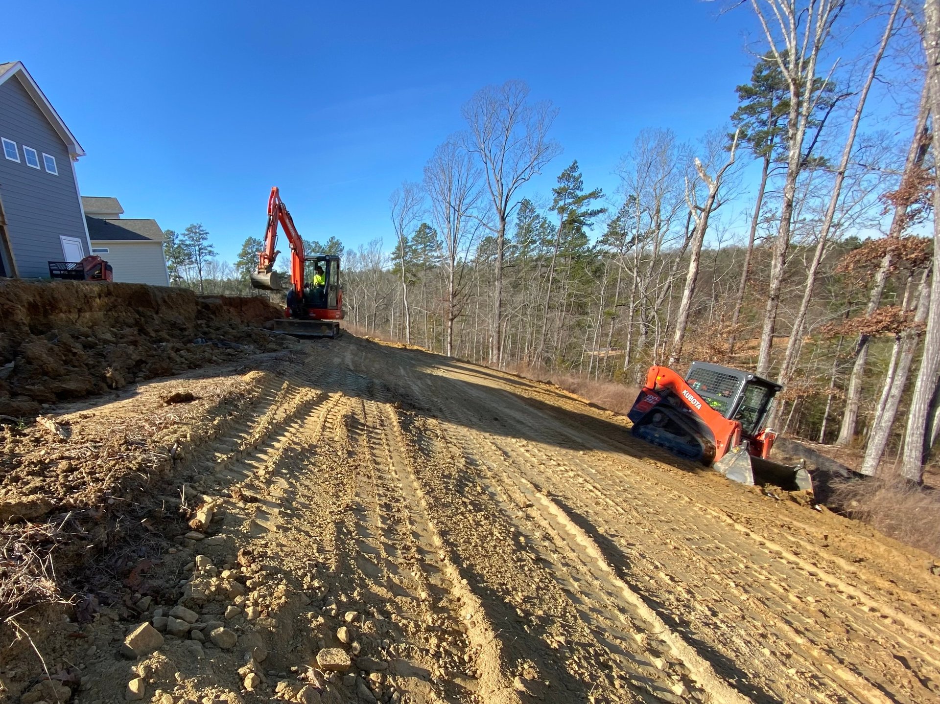 Construction site with excavator and orange machinery on cleared dirt lot, bare trees in background, residential house visible on left