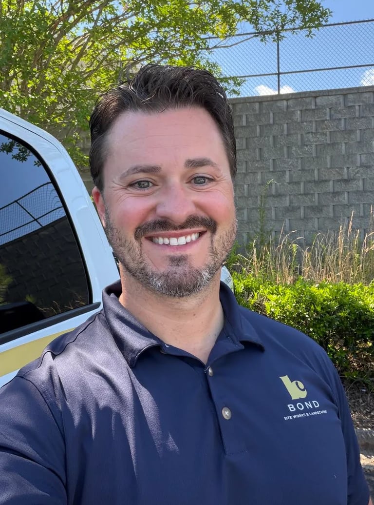Man smiling at camera wearing navy blue BOND polo shirt, standing in front of white vehicle and residential background