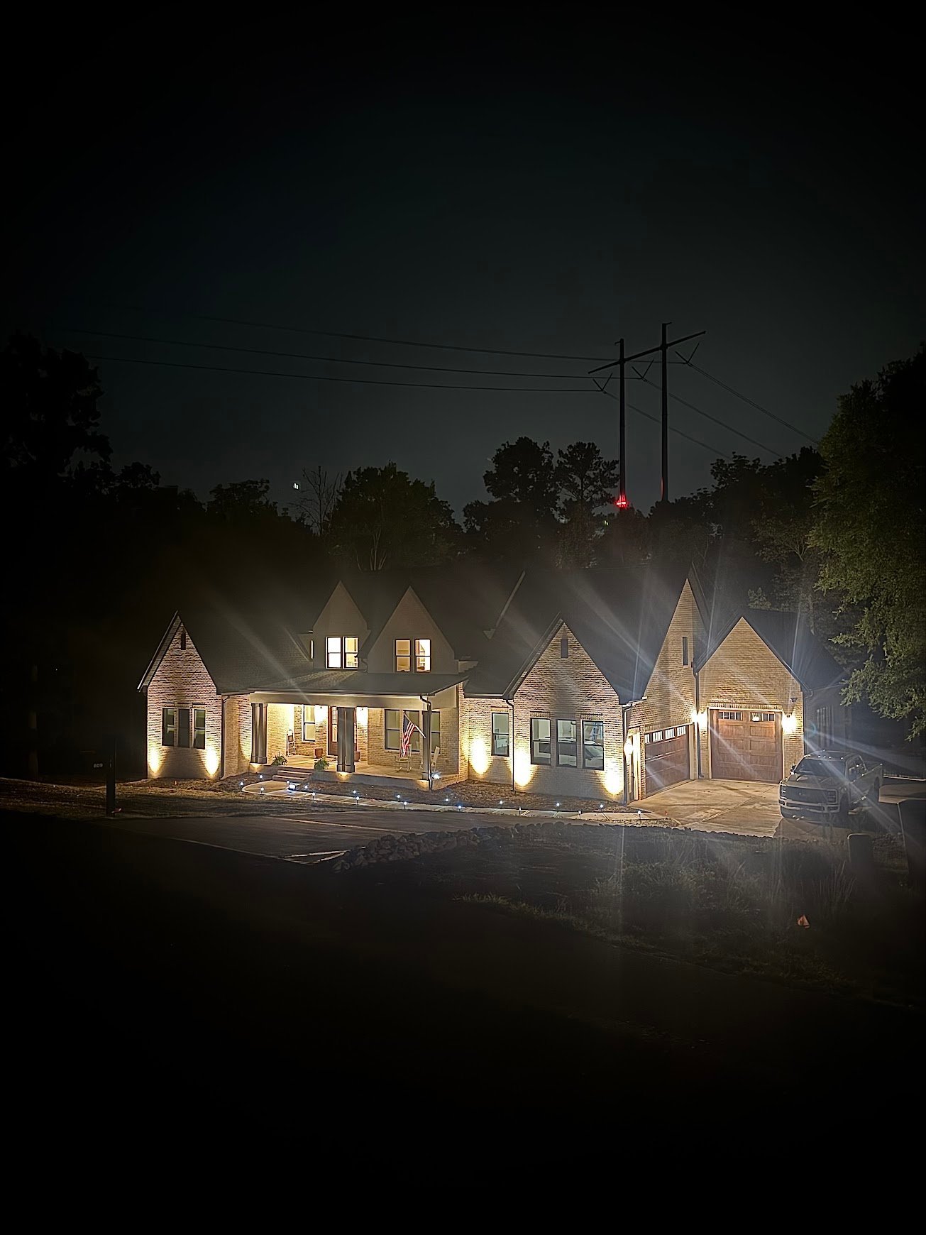 Illuminated residential house with attached garage at night, surrounded by dark trees and power lines overhead