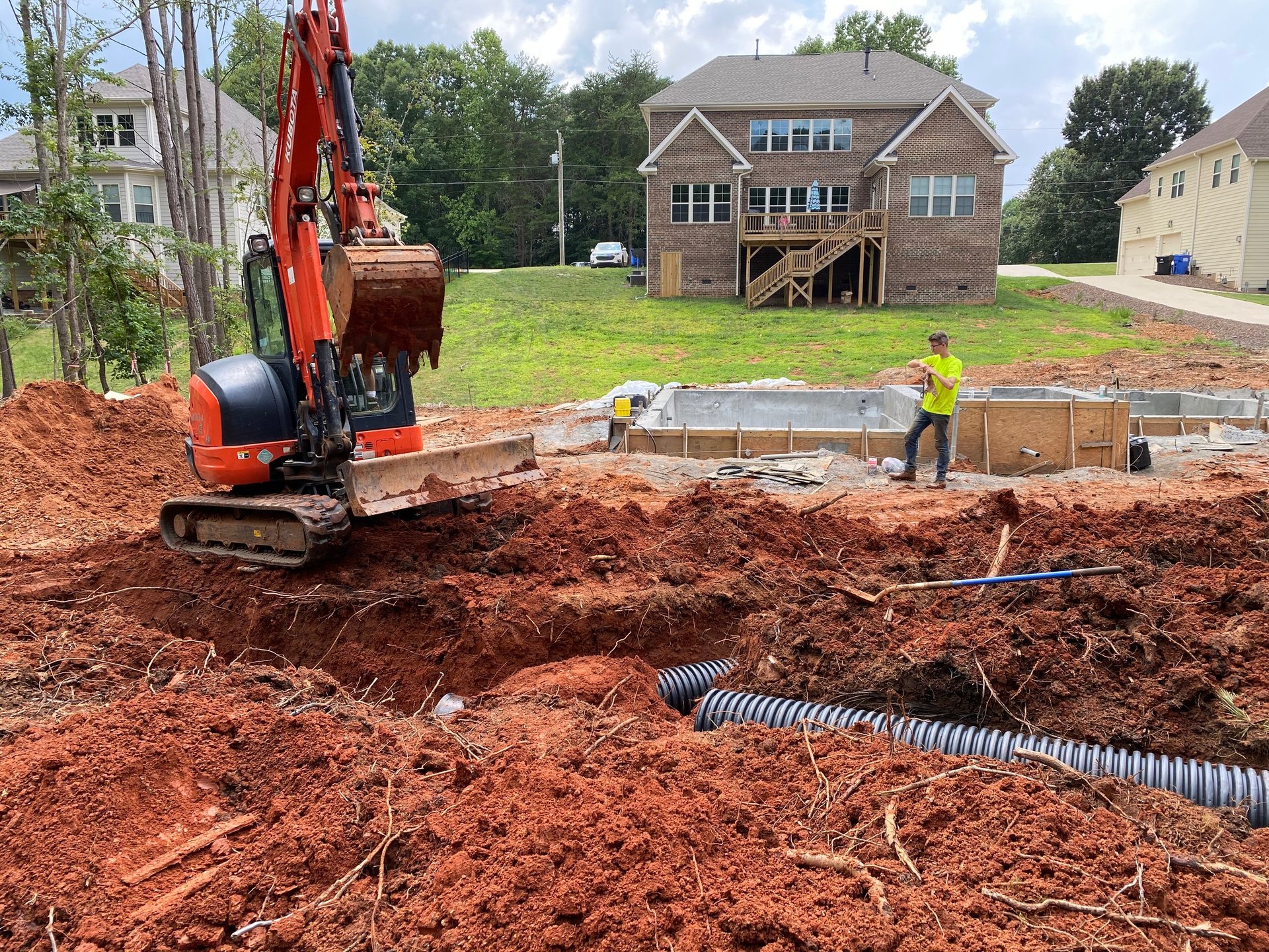 Red mini excavator digging on residential construction site with corrugated drainage pipe visible and brick house in background